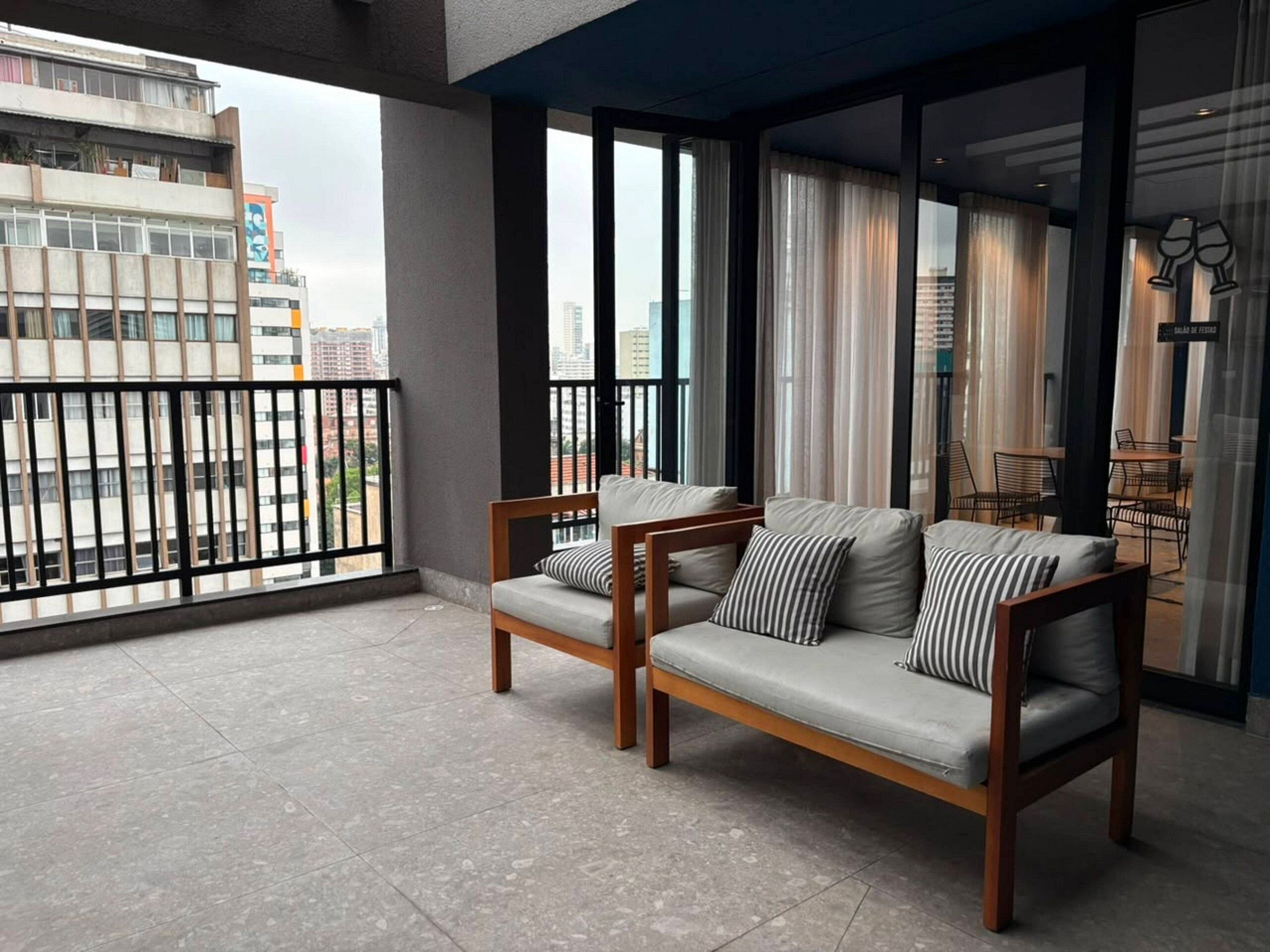 Varanda com dois sofás e uma mesa de jantar, vista para o horizonte urbano e uma placa com o nome do quarto.