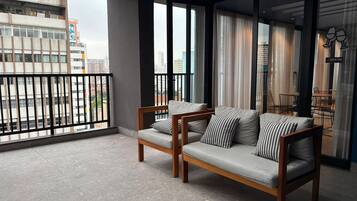 Varanda com dois sofás e uma mesa de jantar, vista para o horizonte urbano e uma placa com o nome do quarto.