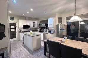 A modern kitchen with white cabinetry, a central island, and a dining area with a table and chairs. - Two bedroom suite in a House/ 15 min YVR, AC (Richmond)