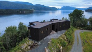 A modern cabin with a sloped roof, situated on a rocky outcrop by a lake with mountains in the background.