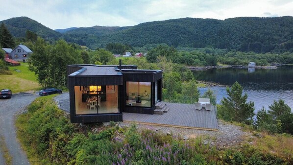 A modern house with a glass facade, a deck, and a view of a lake and forested hills.