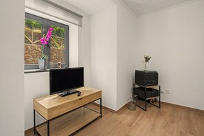 A modern living room with a flat-screen TV on a wooden TV stand, a small black speaker on a shelf, and a window with a view of greenery. - Casa Da Costa: Sea View, Private Terrace & Wi-Fi in São Vicente, Madeira (Sao Vicente)