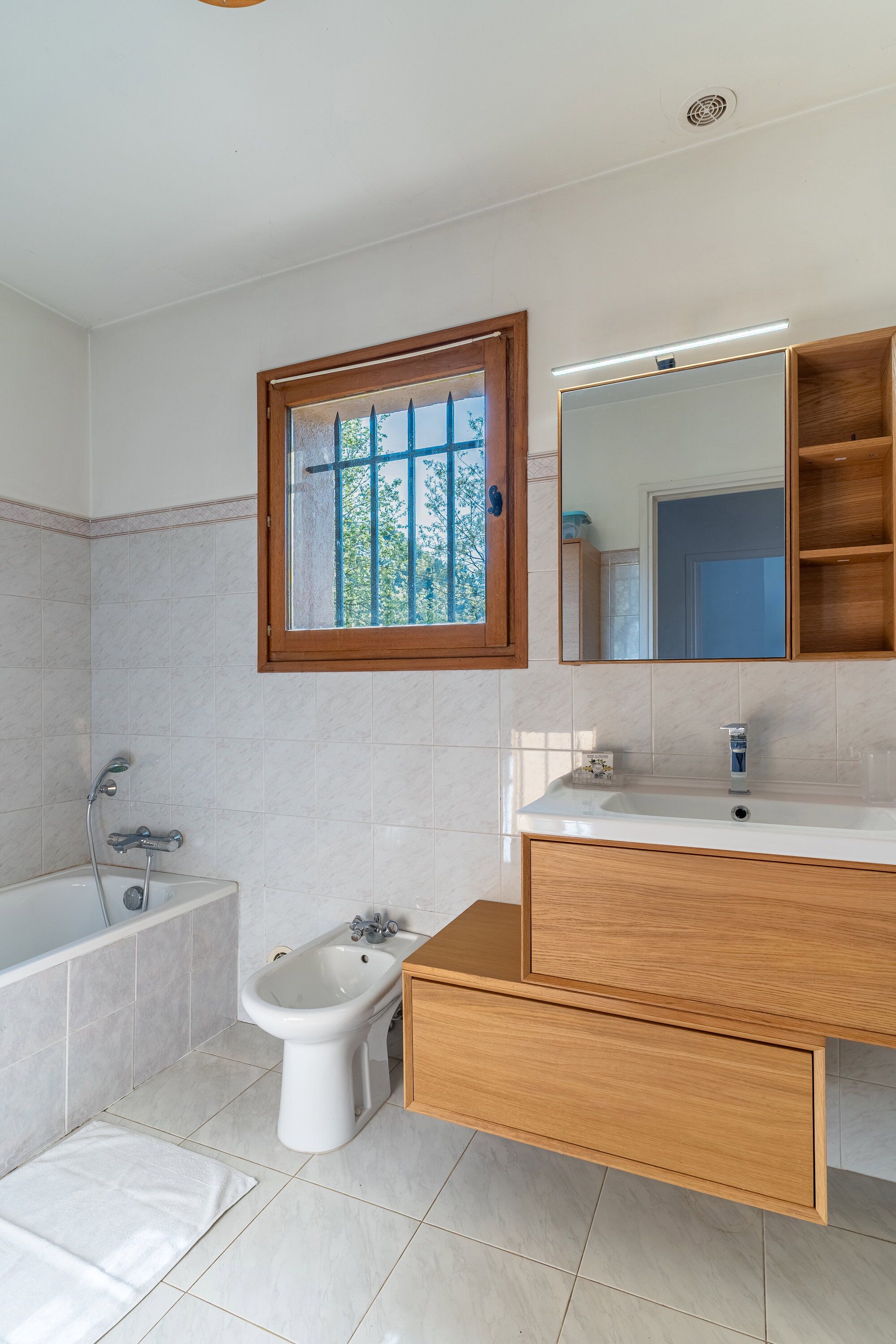 A modern bathroom with a bathtub, bidet, vanity with mirror, and a window with a view of trees.