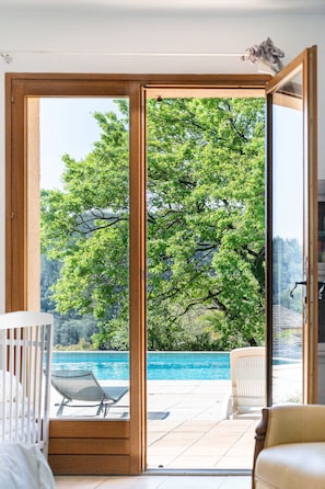 A room with a large sliding glass door leading to a poolside area with a view of a tree and a body of water. - Écrin de verdure, charme provençal près de Cannes (Mouans-Sartoux)