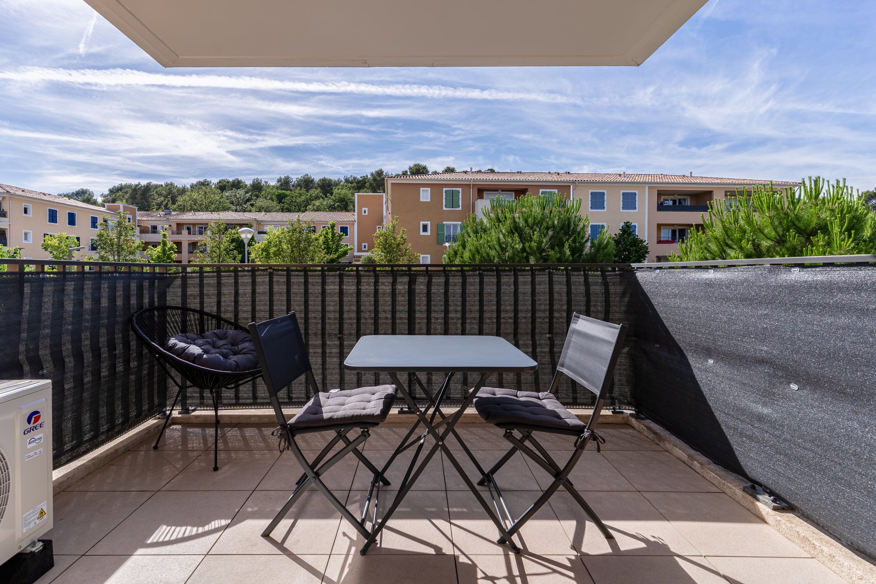 A terrace with a table and two chairs, a black chair with a cushion, and a white air conditioning unit.