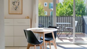 A modern dining area with a round table, two chairs, and a view of a balcony with a railing and greenery. - Escale Calistea : tout confort avec piscine & clim (Roquefort-les-Pins)