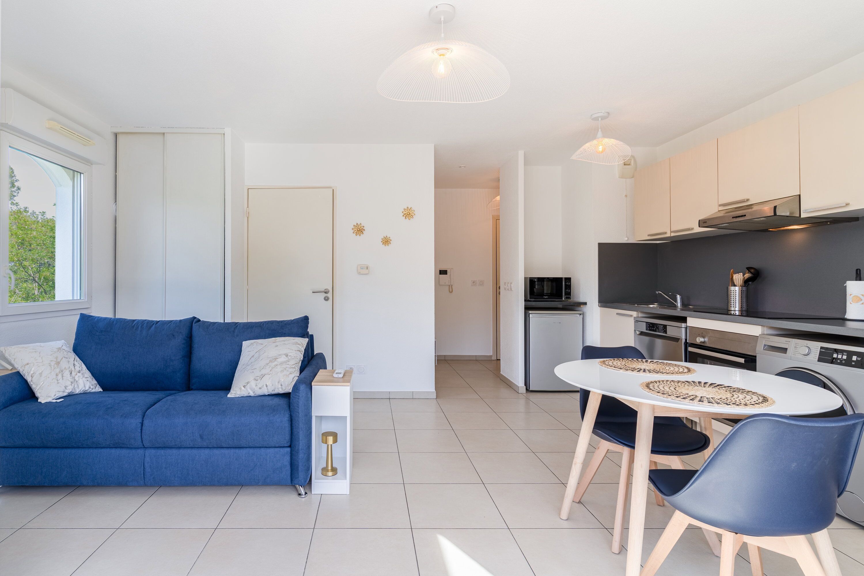 Ein modernes Apartment mit einem blauen Sofa, einem Esstisch mit Stühlen, einer Küchenzeile und einem großen Fenster.
