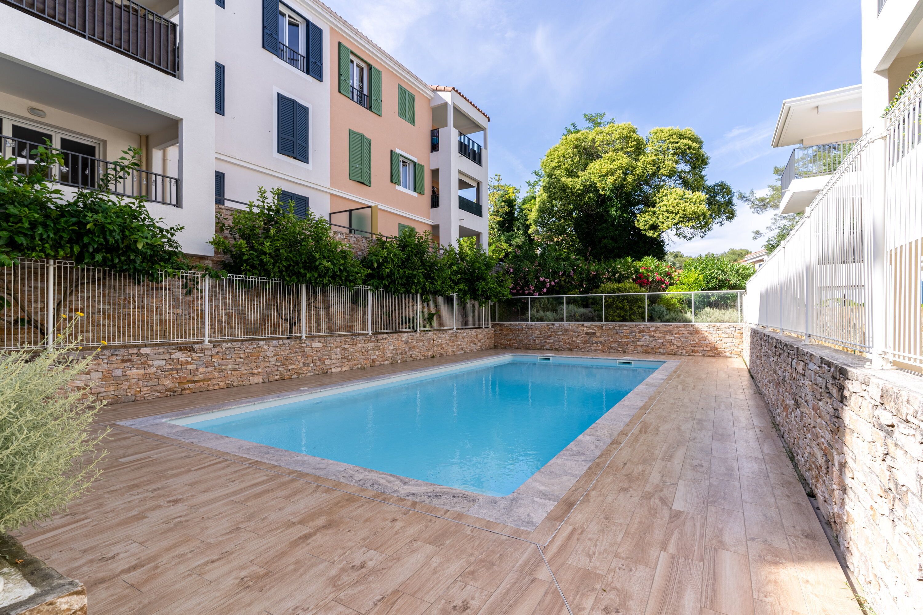 Une piscine rectangulaire bordée d’un mur en pierre et dotée d’une terrasse en bois, le tout entouré d’une clôture en métal et d’une végétation luxuriante.