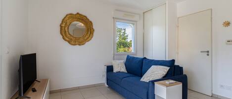 A room with a blue sofa, a television on a stand, a gold-framed mirror, and a window with a view of greenery.