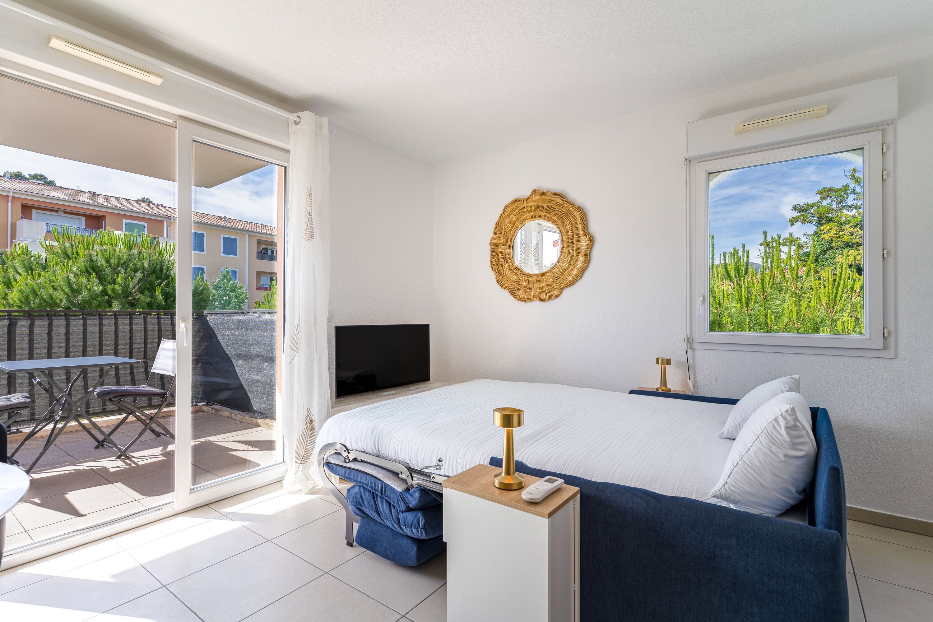 Een slaapkamer met een bed, een televisie, een nachtkastje met een lamp, een raam met uitzicht en een glazen schuifdeur die leidt naar een terras met een tafel en stoelen.