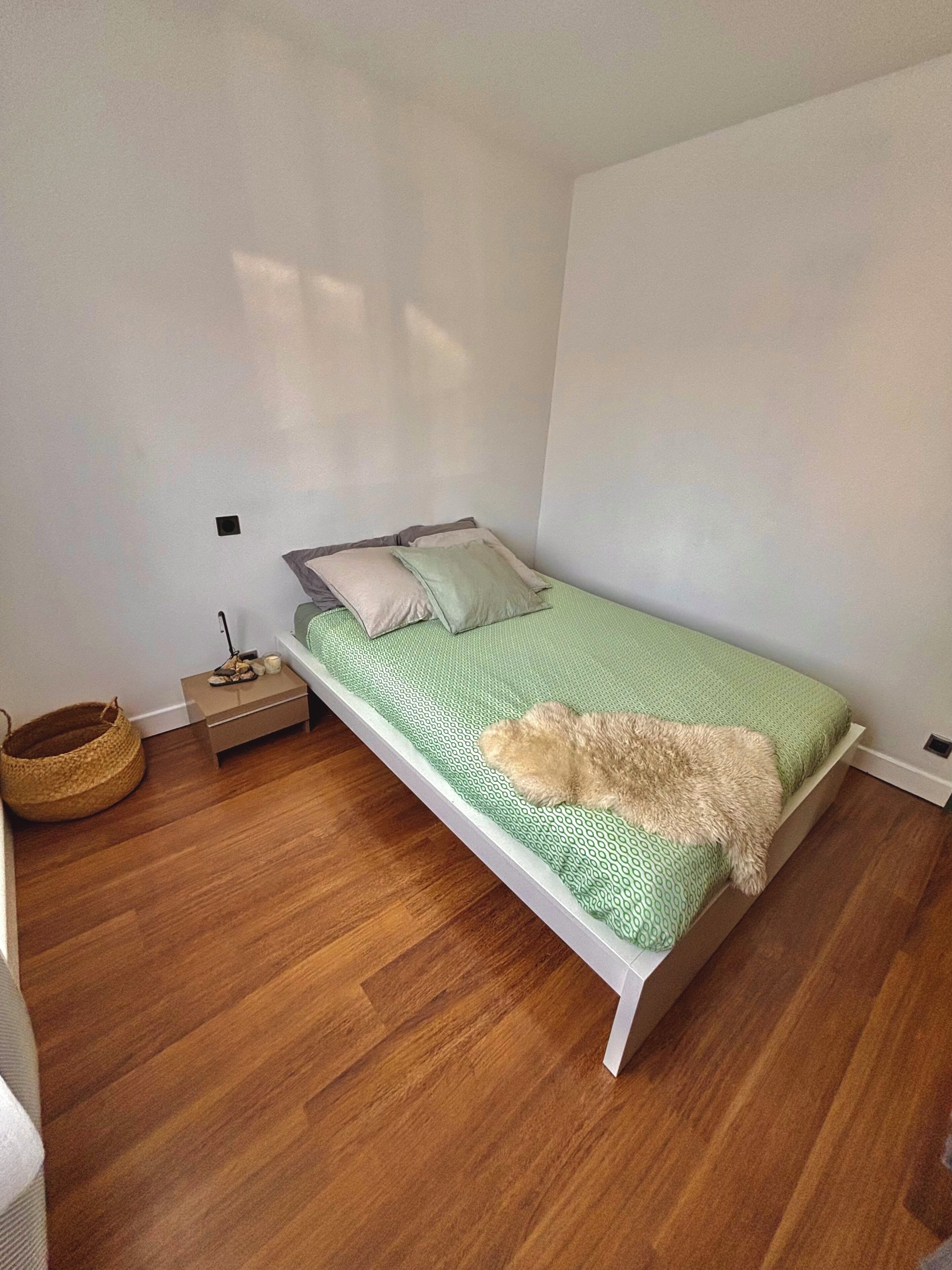 Habitación individual con piso de madera, colcha estampada verde y una mesita de noche con lámpara.