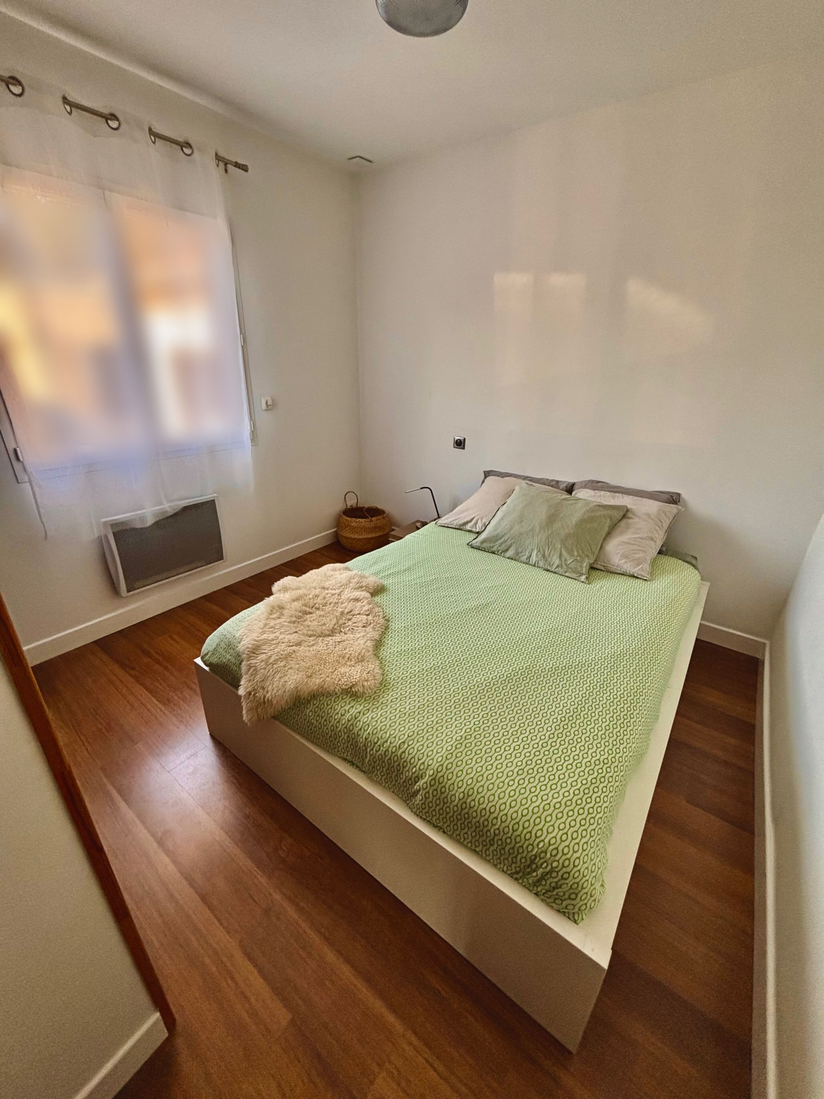 A bedroom with a bed, a window with curtains, a small bedside table, and a wall-mounted heater.