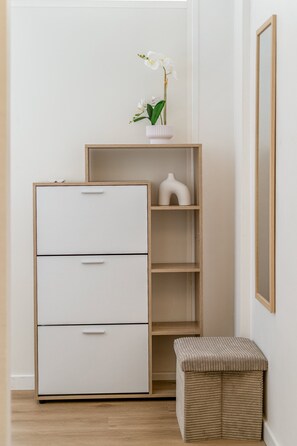 Ein schlichter Raum mit einem weißen Aktenschrank, einem hölzernen Bücherregal und einer gewebten Bank.