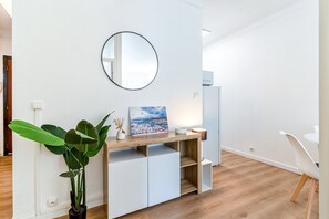 Ein modernes Zimmer mit weißer Wand, einem runden Spiegel, einem Holztisch mit einem Bild, einer Topfpflanze und einem weißen Stuhl.