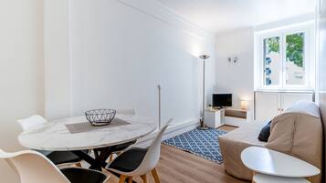 Una sala de estar moderna y minimalista con una mesa redonda de comedor, sillas blancas, un sofá y un televisor.
