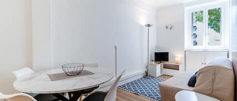 Una sala de estar moderna y minimalista con una mesa redonda de comedor, sillas blancas, un sofá y un televisor.