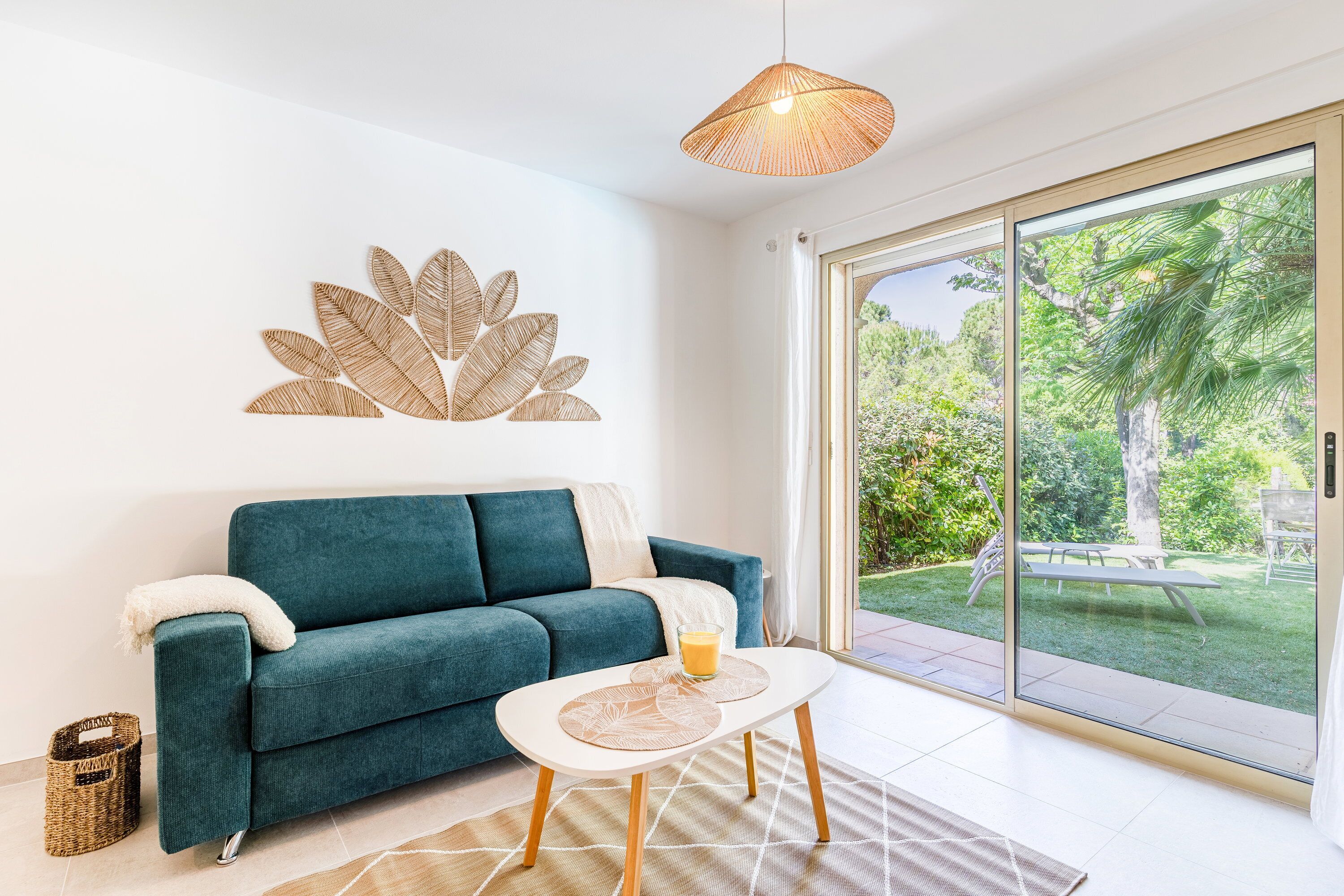A modern living room with a teal sofa, a glass coffee table, and a large sliding glass door leading to a garden.