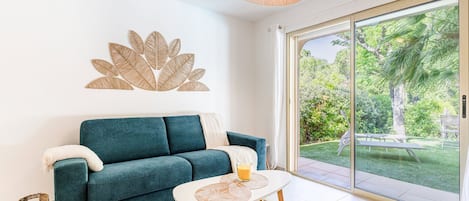 A modern living room with a teal sofa, a glass coffee table, and a large sliding glass door leading to a garden.