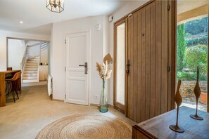 A spacious room with a wooden door, a staircase leading to another level, a dining area with a table and chairs, and a view of a garden through a window. - La Villa Estérelle — Vue golf, piscine & élégance (Saint Raphael)
