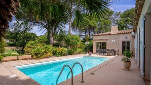 A swimming pool with a patio area, surrounded by lush greenery and a building with a tiled roof. - La Villa Estérelle — Vue golf, piscine & élégance (Saint Raphael)