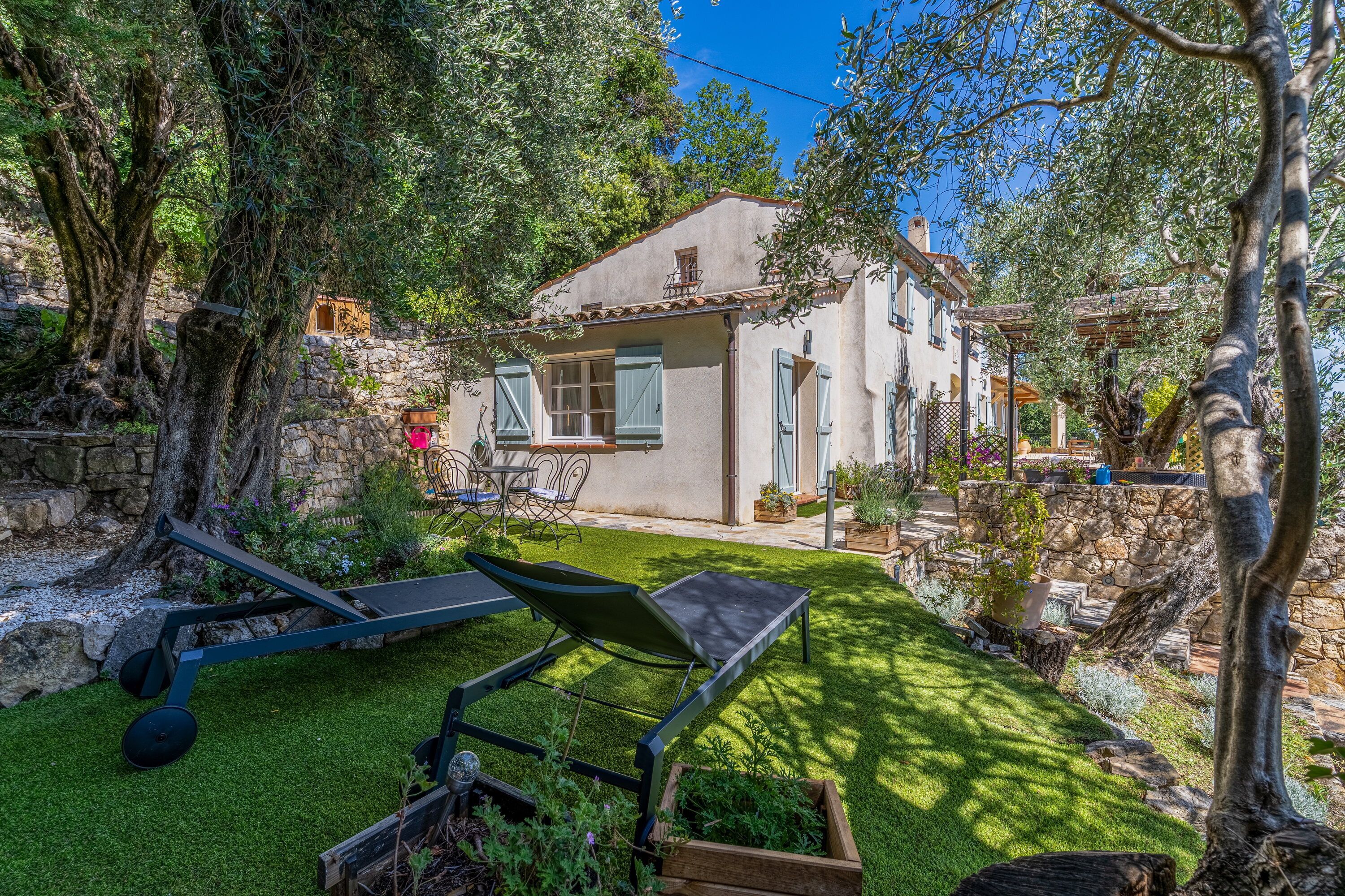 Une maison avec un mur en pierre, une terrasse avec une table et des chaises, et une pelouse.