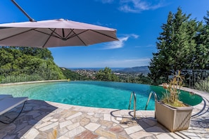 A swimming pool with a stone-tiled deck, a metal pool ladder, and a large umbrella. - L'Échappée Parfumée, Panorama d'exception et calme (Grasse)