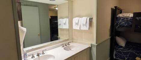 A bathroom with a double sink, a large mirror, and a bed with a blue patterned bedspread.