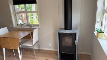 Une pièce avec une table en bois, une chaise blanche, une fenêtre avec des stores et un poêle à bois noir.