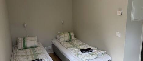 A small, single-room accommodation with two single beds, a bedside table, and a power outlet on the wall.