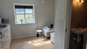 Une petite pièce comprenant un lit, un petit tabouret, une fenêtre avec des stores, un lavabo blanc et un four à micro-ondes.