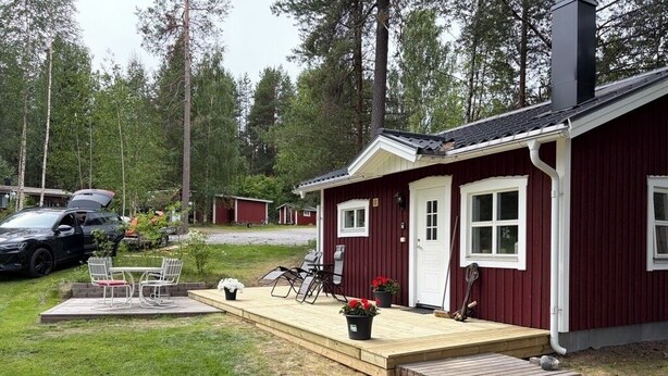 Un bâtiment rouge avec un porche, une voiture garée devant et un espace verdoyant aménagé avec du mobilier d’extérieur.