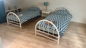 Une chambre à coucher avec deux lits à structure métallique, une table de chevet avec une lampe et une étagère contenant des livres et des photos encadrées.