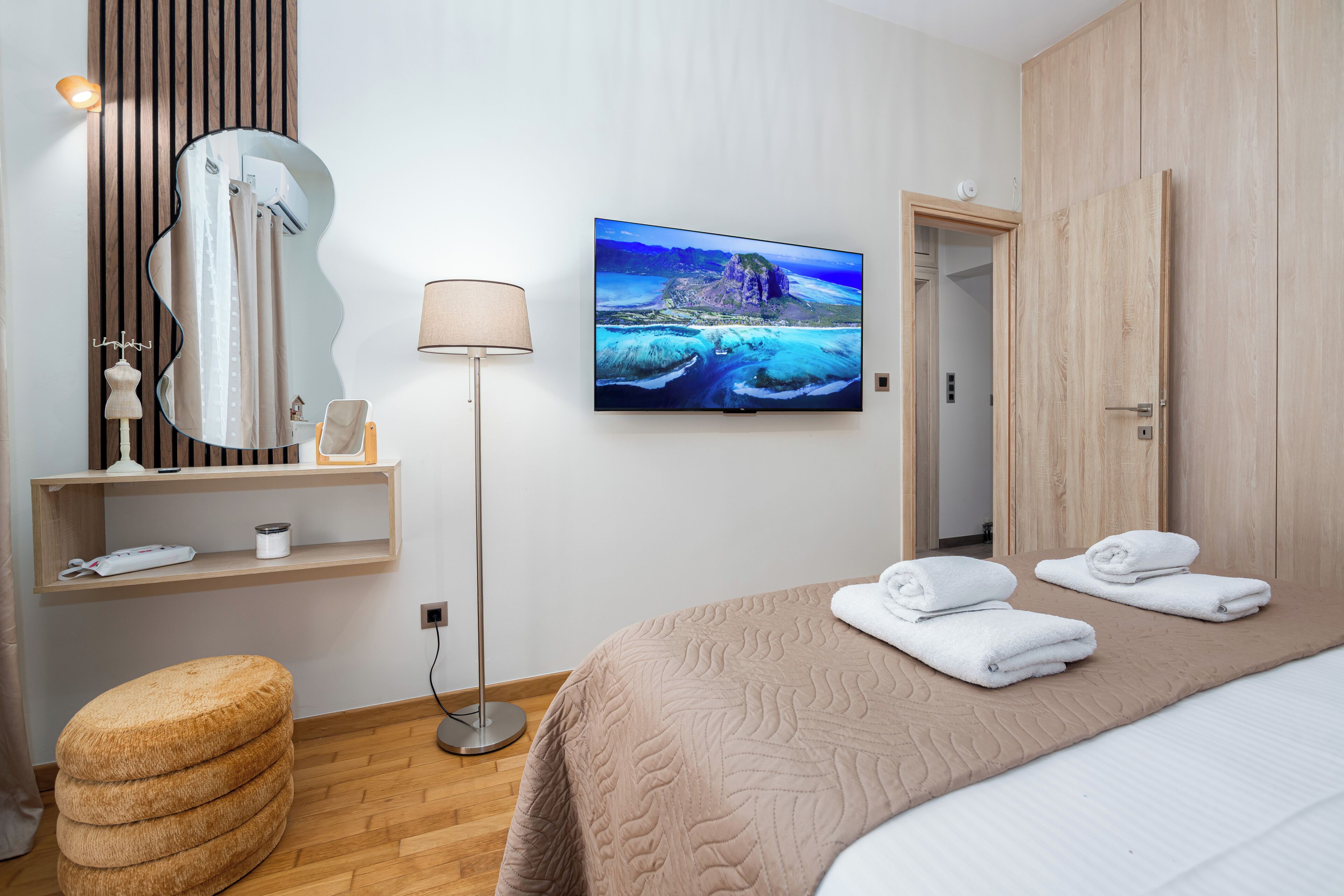 Une chambre d’hôtel comprenant un lit, un téléviseur à écran plat, un miroir, une lampe, une armoire en bois et un petit tabouret.