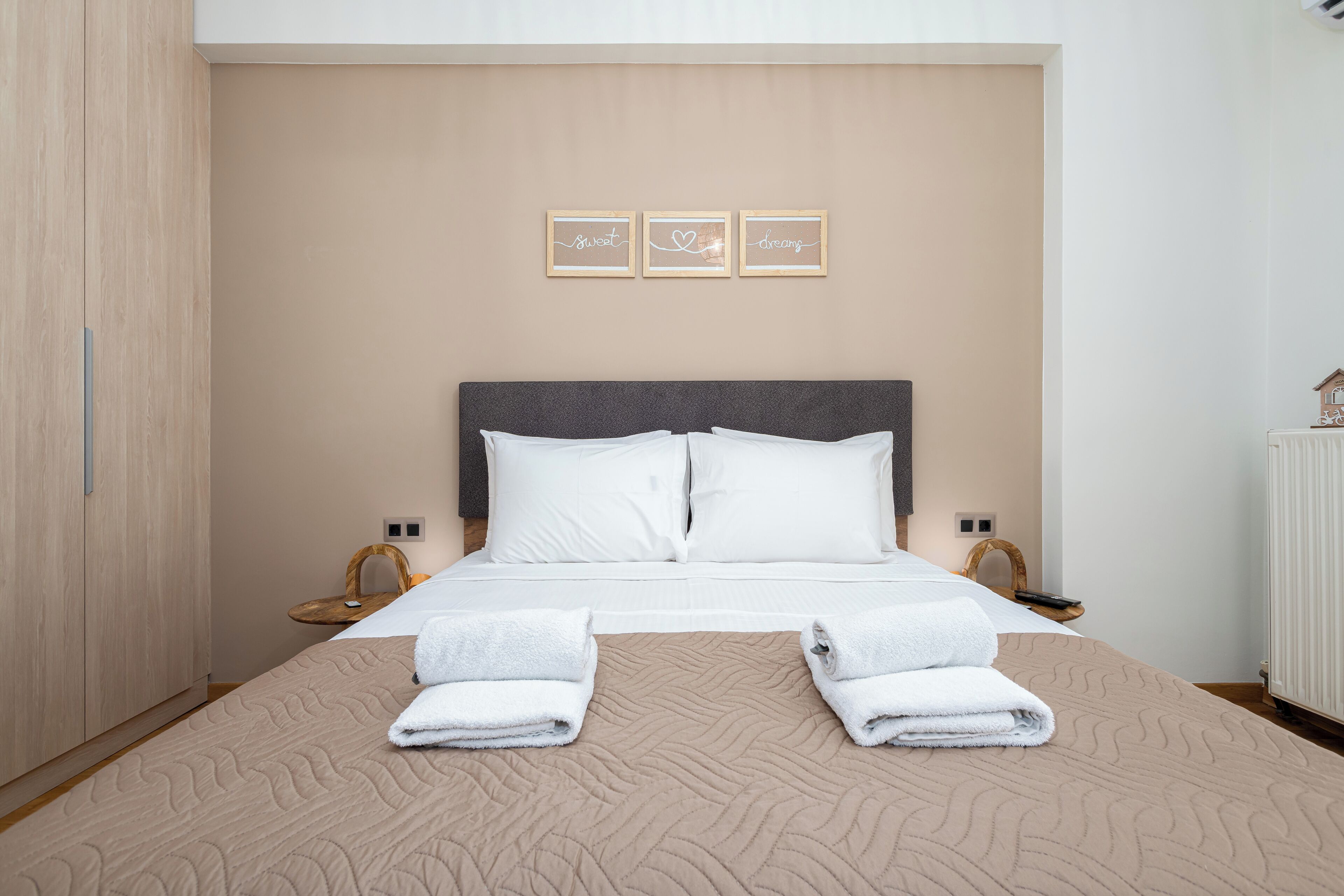 A neatly made bed with white linens and two folded towels, a grey headboard, and three framed wall art pieces with text.