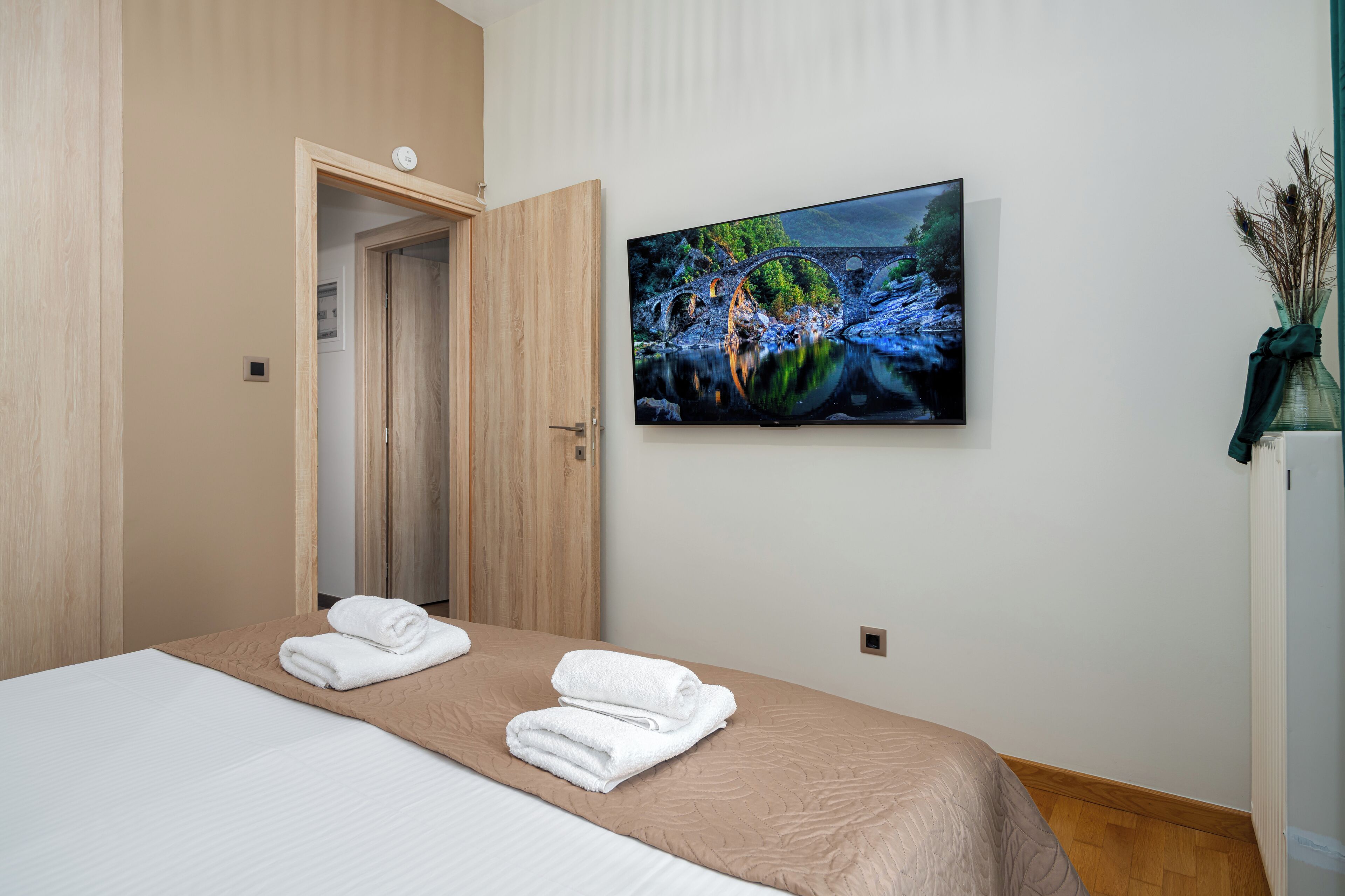 A hotel room with a bed, a painting of a bridge over water, and a vase with dried plants.