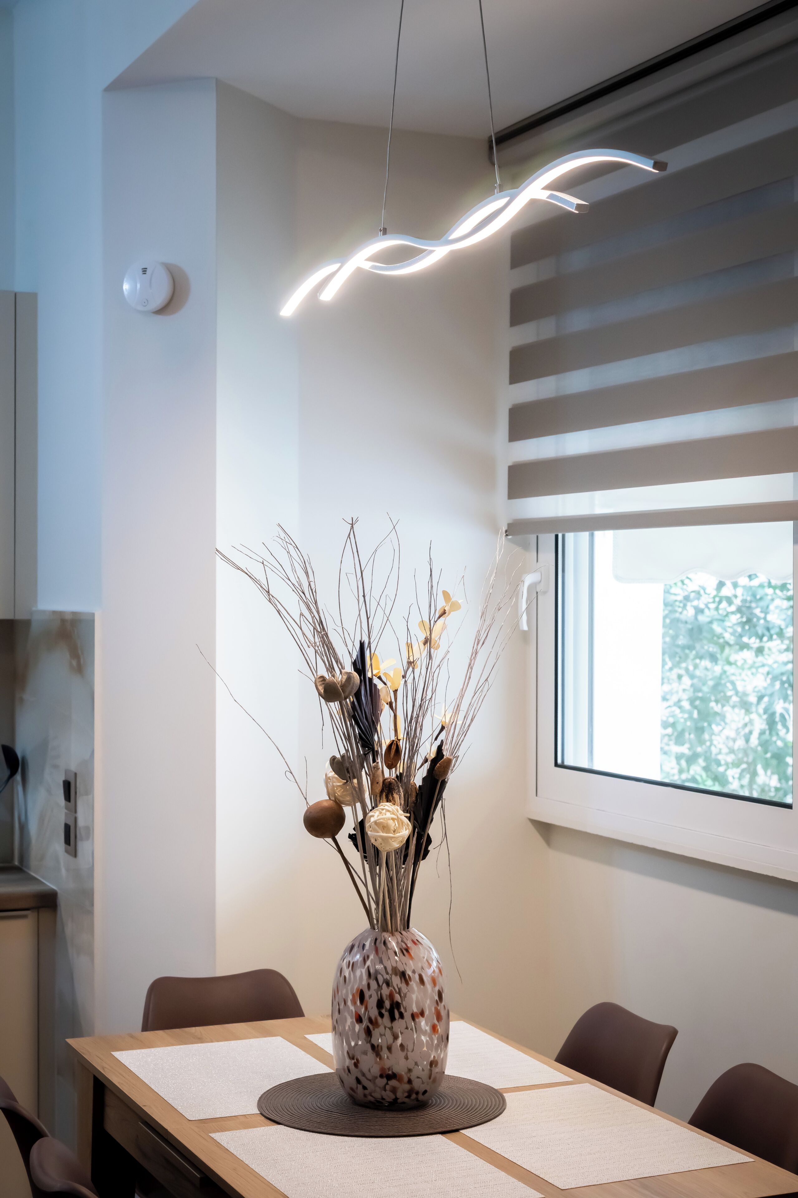 A dining area with a table, chairs, a vase with dried plants, a hanging light fixture, and a window with blinds.