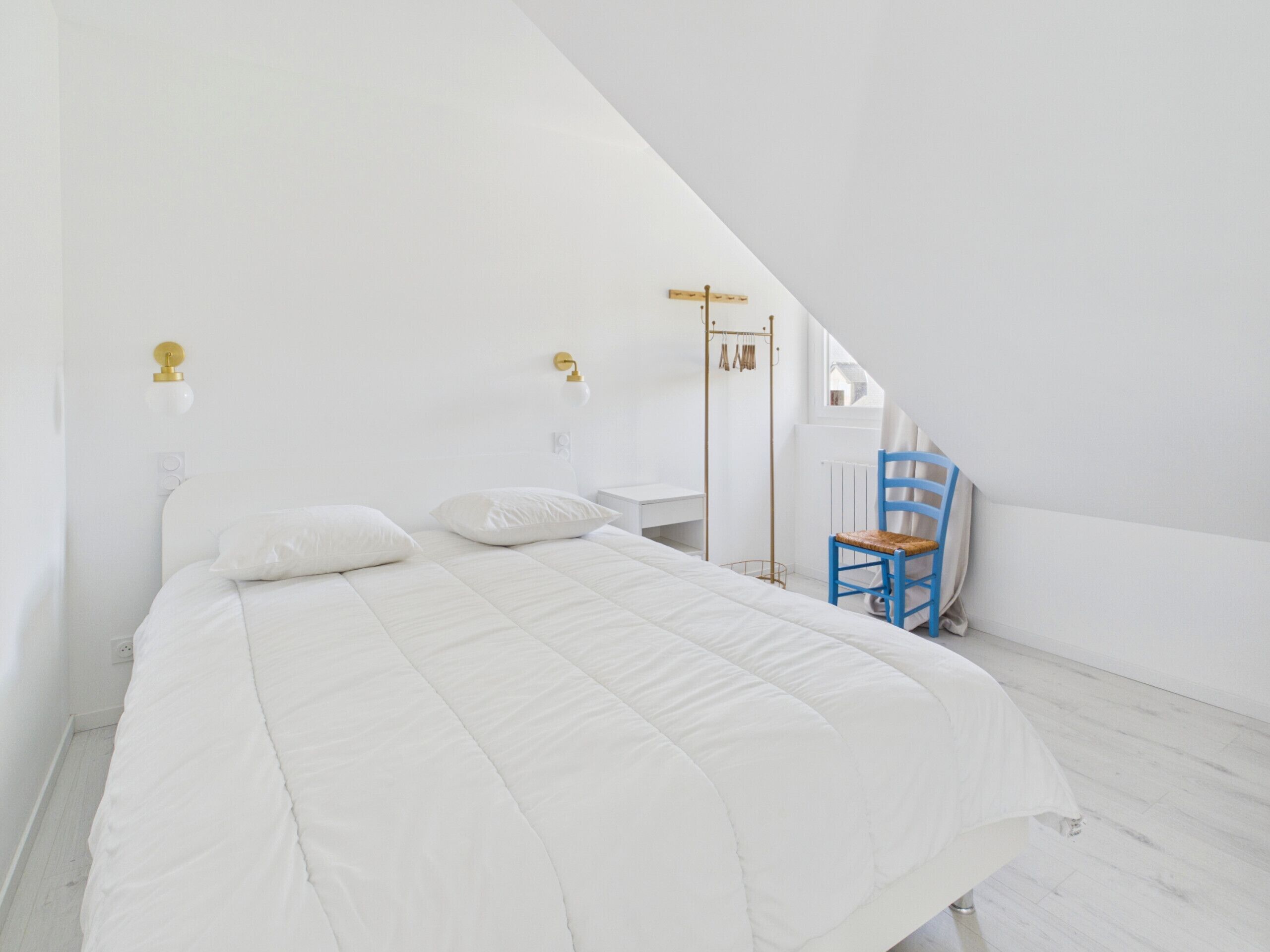 A white bedroom with a bed, two wall-mounted lamps, a blue chair, and a radiator.