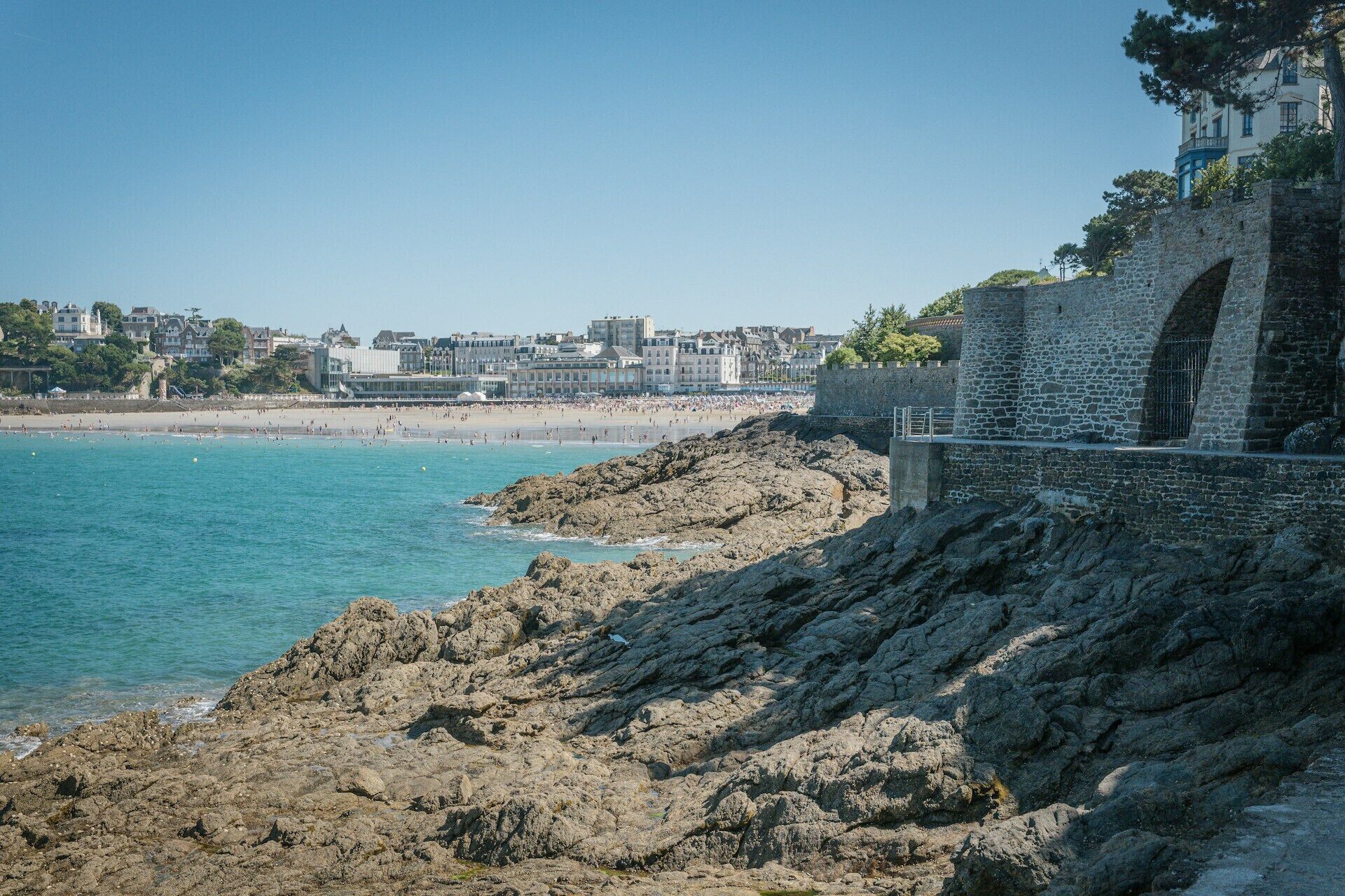 A coastal view with a rocky shoreline, a stone bridge, and buildings in the background.