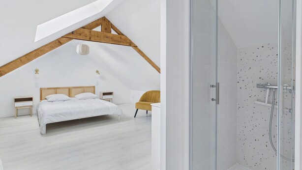 Ein Schlafzimmer mit schrägverdeckter Decke, einem Bett, einem Sessel und einer Duschkabine aus Glas.