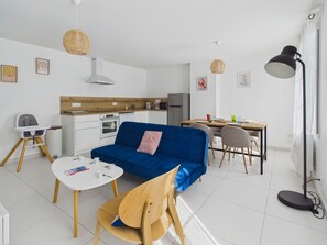 Ein modernes Wohnzimmer mit einem blauen Sofa, Holzstühlen, einem Esstisch und einer kleinen Küchenzeile im Hintergrund.
