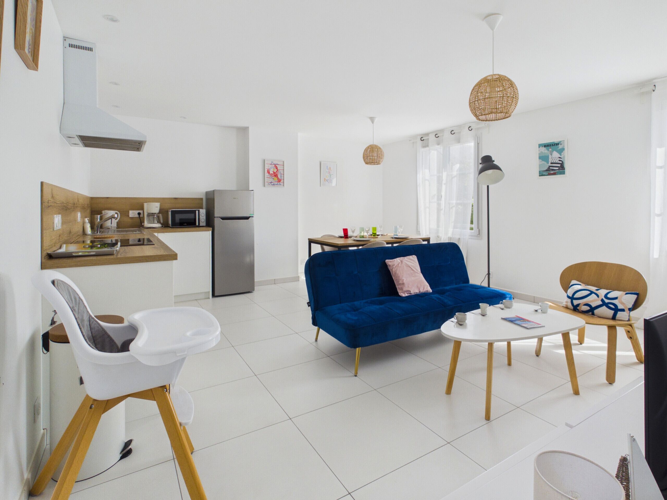 Un salon moderne avec un canapé bleu, un fauteuil blanc, une table à manger avec des chaises, un coin cuisine équipé d’un réfrigérateur et d’un four à micro-ondes, et une grande fenêtre avec des rideaux.