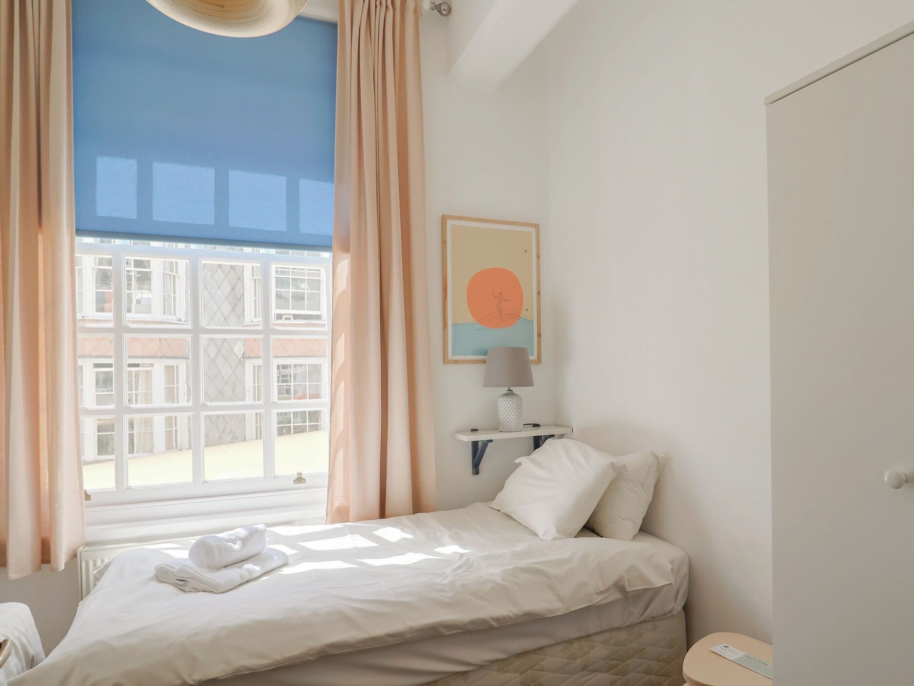Une chambre à coucher avec un lit, une table de chevet avec une lampe, une fenêtre avec des rideaux et une étagère murale avec un tableau.