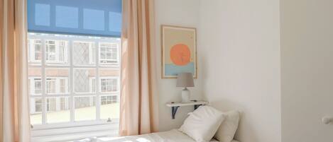 Une chambre à coucher avec un lit, une table de chevet avec une lampe, une fenêtre avec des rideaux et une étagère murale avec un tableau.