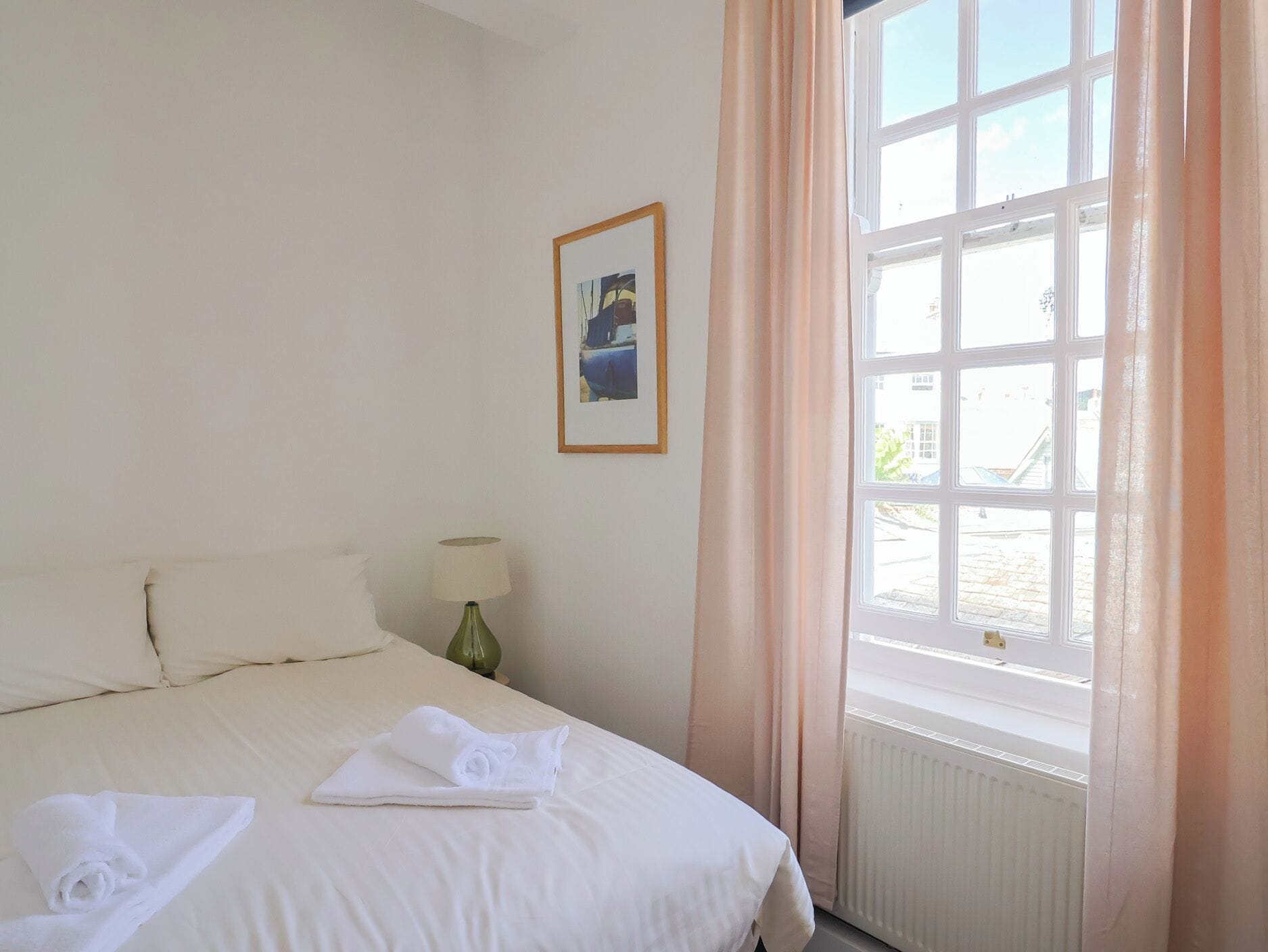 Quarto de hotel com uma cama, mesa de cabeceira com abajur, quadro na parede e janela com cortinas.