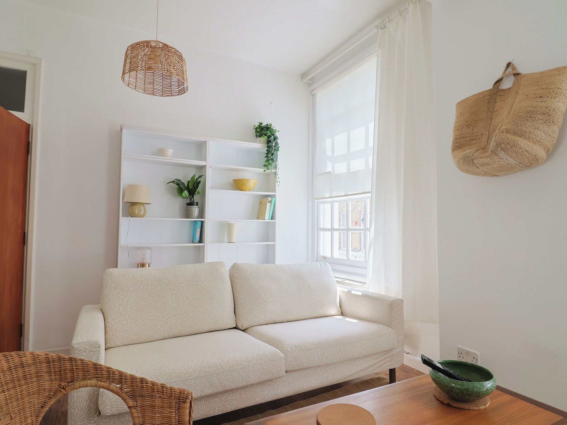A modern living room with a sofa, a wicker chair, a wooden table, a green bowl, and a white shelving unit with various items.