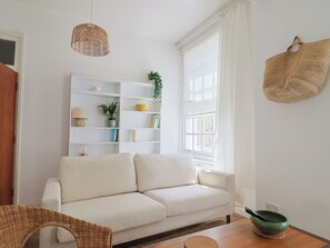 Una sala moderna con un sofá, una silla de mimbre, una mesa de madera, un cuenco verde y una estantería blanca con varios objetos.
