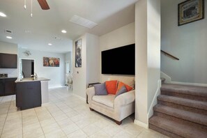 Ein Wohnzimmer mit einer Couch, einem flachbildfernseher an der Wand, einer Küchenzeile mit Kühlschrank und einer Treppe.