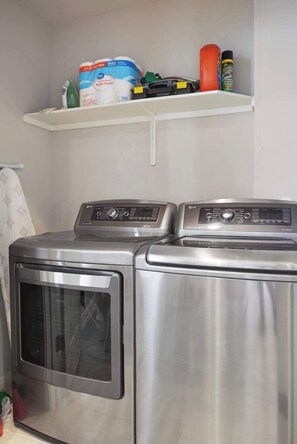 A laundry area with two stainless steel front-loading washers and dryers, a shelf above containing cleaning supplies, and a towel rack to the left. - Modern 2BR Haven near Austin & Tesla (Manor)