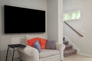Ein Wohnzimmer mit einem an der Wand montierten Flachbildfernseher, einem weißen Sofa mit bunten Kissen und einer Holztreppe mit einem Fenster darüber.