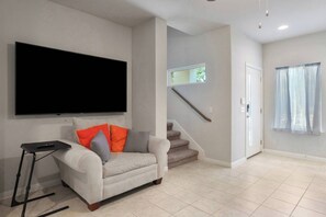 A living room with a flat-screen TV, a sofa with cushions, a remote control, and a staircase leading to another room. - Modern 2BR Haven near Austin & Tesla (Manor)